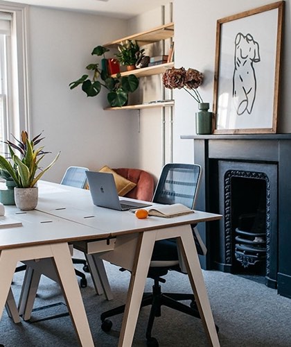 Bright studio workspace with shared desks, plants, and a framed drawing above the fireplace.